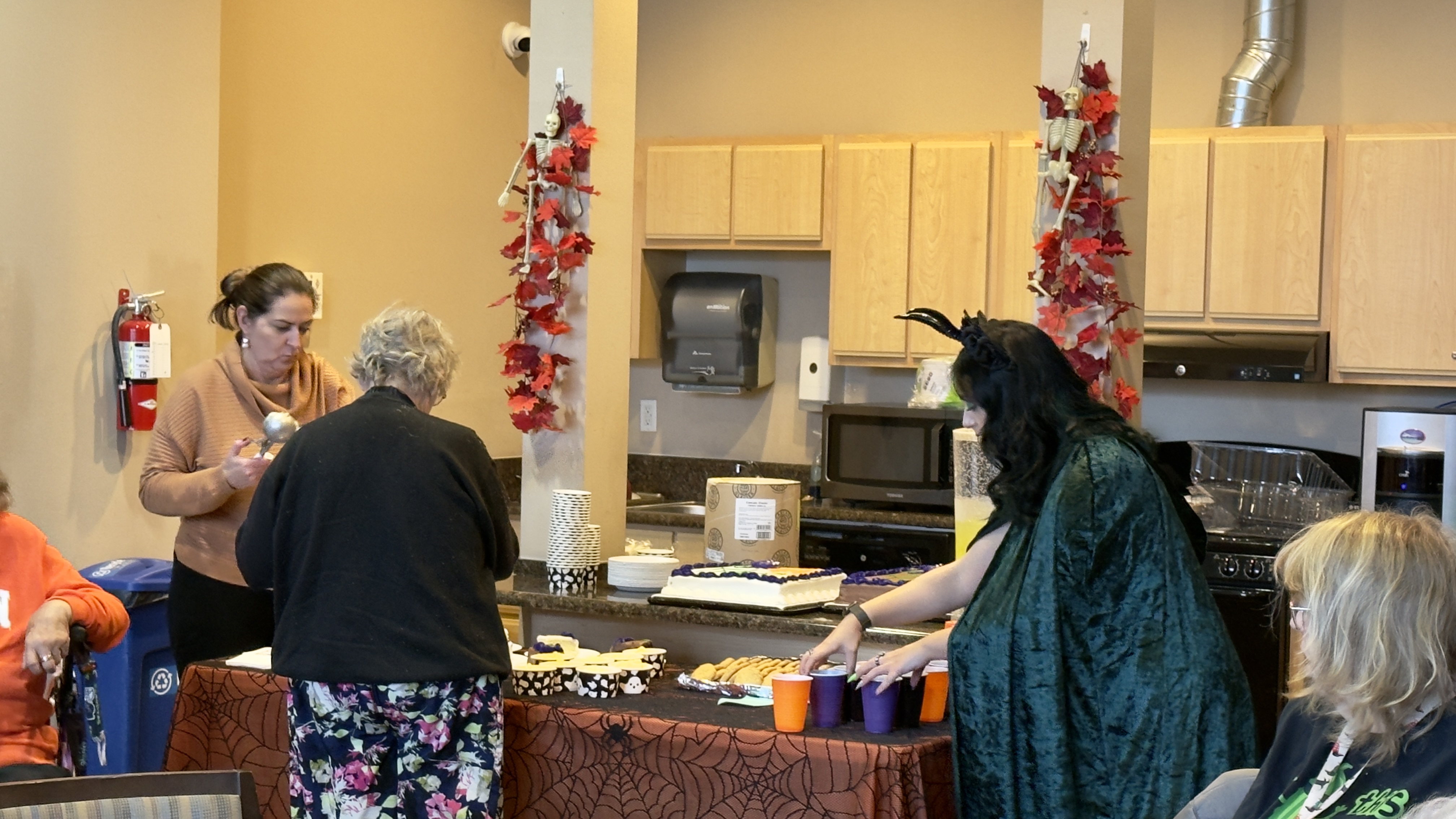 Two women serve desserts and drinks at a Halloween-themed table decorated with spiderweb cloth and fall leaves.